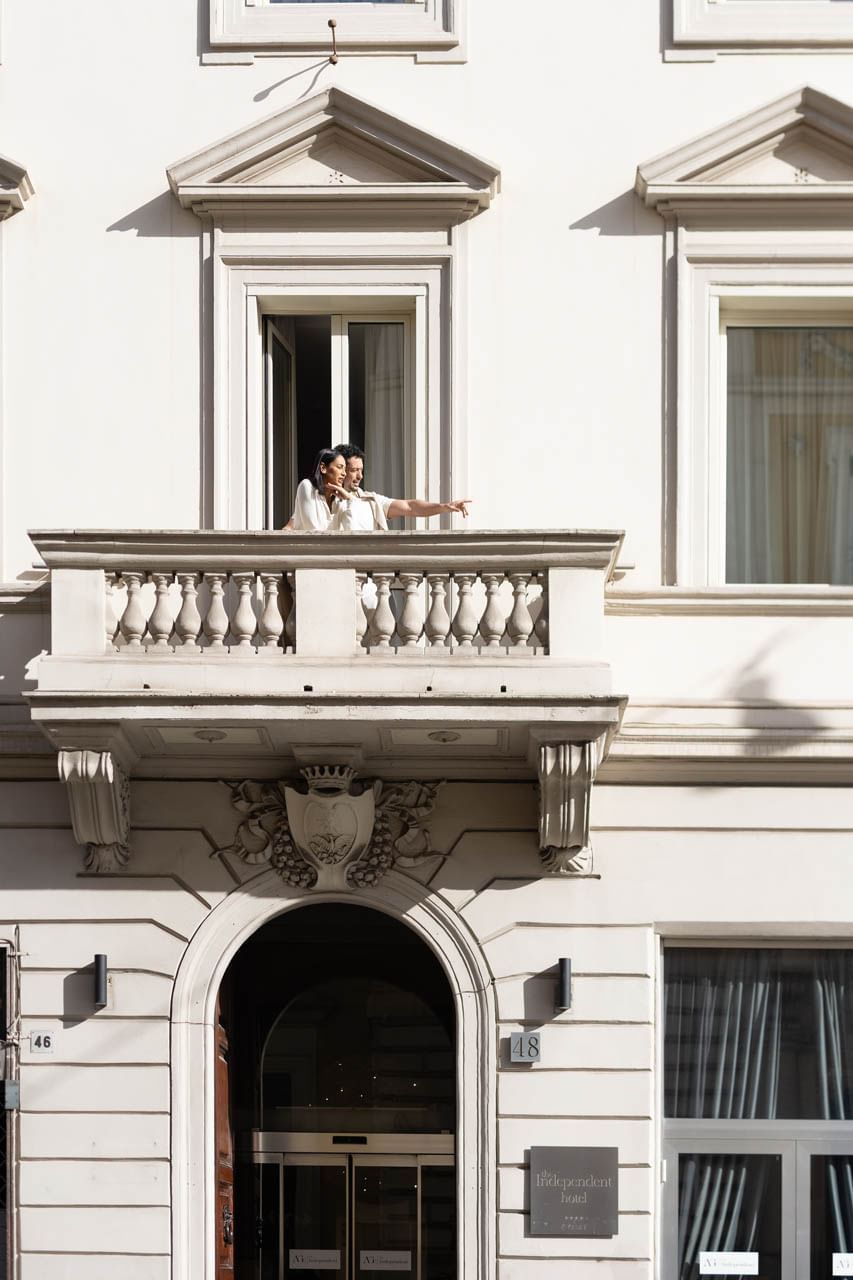 A couple stands on a balcony at The Independent, gesturing toward the street with ornate architecture