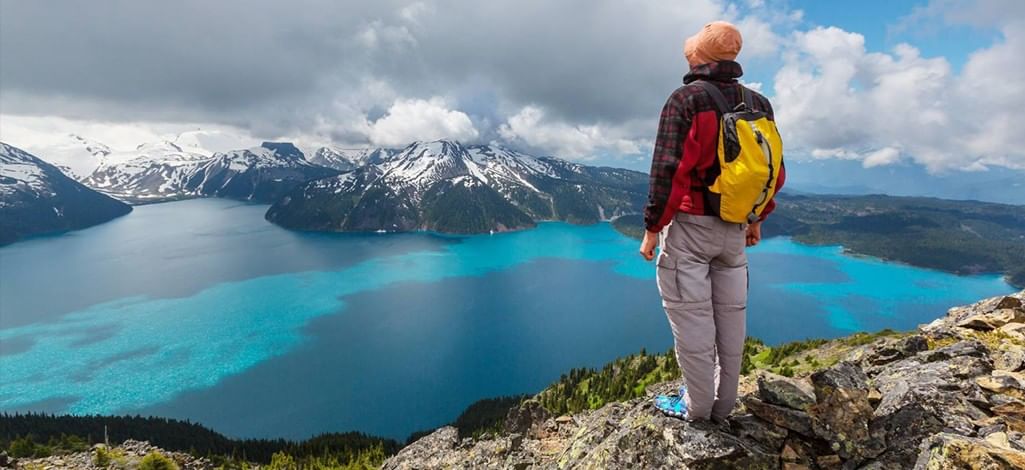 Person standing on a rocky mountain summit wearing outdoor clothing and a backpack, overlooking a turquoise alpine lake surrounded by snow-capped peaks.