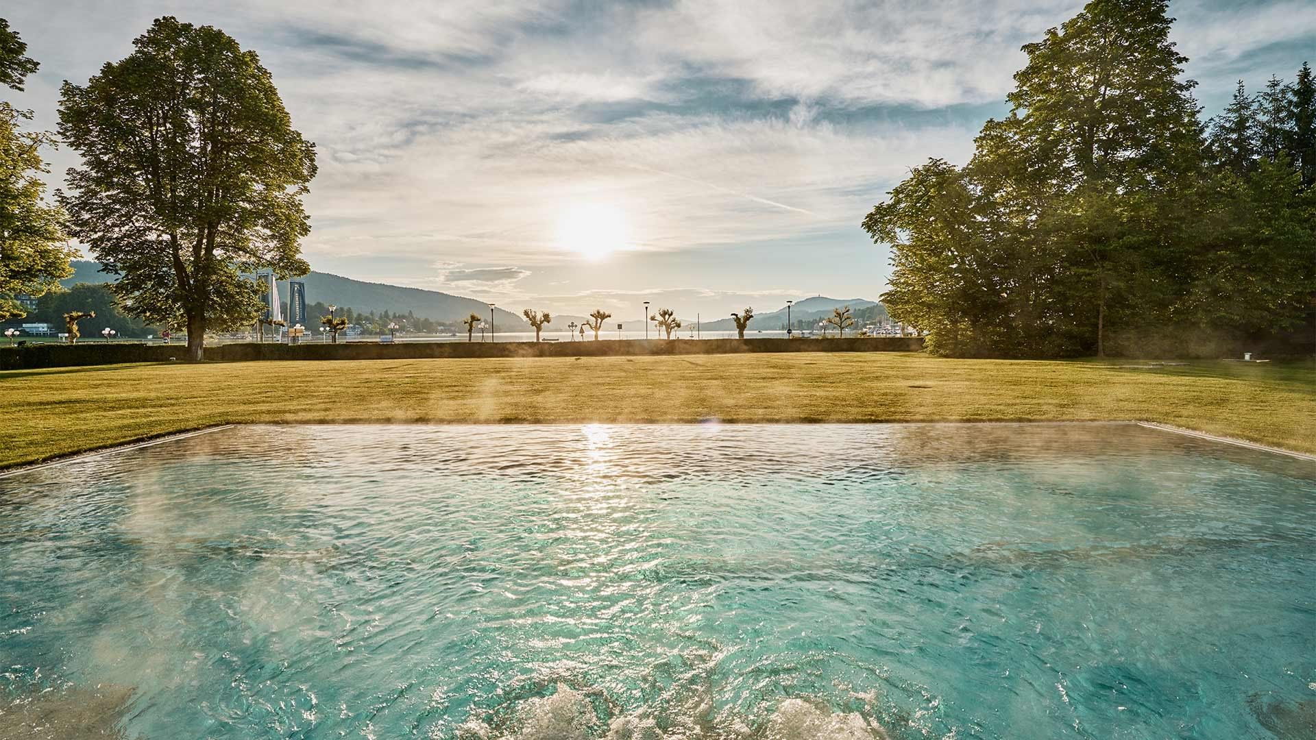 Outdoor pool with blue water, surrounded by trees and a grassy area, with mountains in the background.