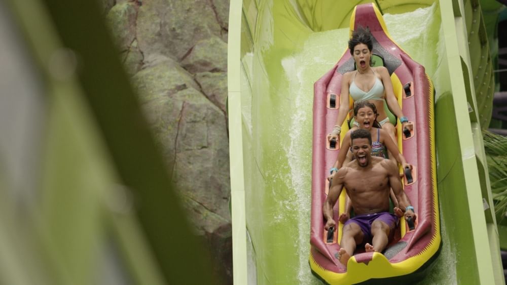 Three people screaming in excitement and holding onto a red and yellow raft as they zoom down a green waterslide. 