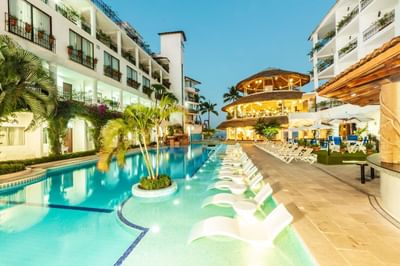 Illuminated pool with white lounge chairs surrounded by white hotel buildings and palm trees at Playa Los Arcos Puerto Vallarta