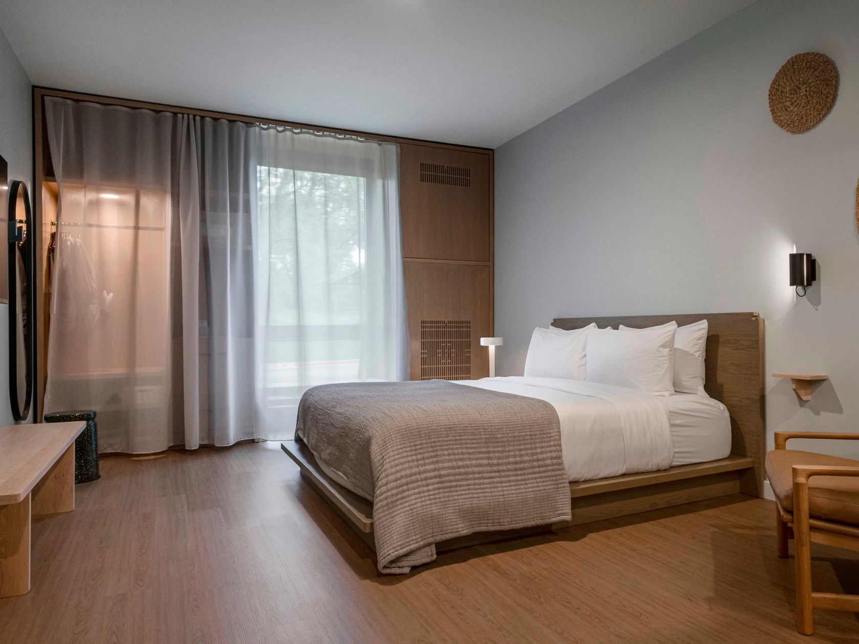 Modern bedroom with wooden floor, large bed, chair, nightstand, white curtains, and decorative wall elements.