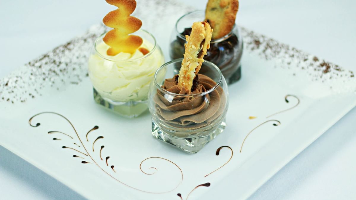 Trois verrines de desserts variés sur une assiette décorée de motifs dorés et de poudre de cacao.