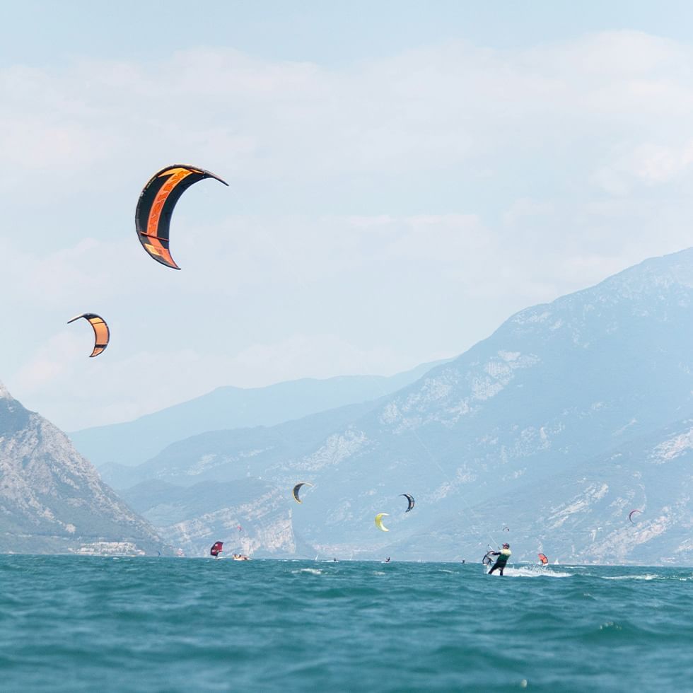 Molti windsurfer navigano nelle acque blu sotto le montagne, con paracadute arancioni e neri che spiccano.