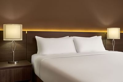 Close up of guest room bed with two pillows and two lamps on a brown nightstand against a brown wall.