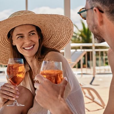 Zwei Personen mit Weingläsern sitzend neben einem Pool, die Frau trägt einen Strohhut.