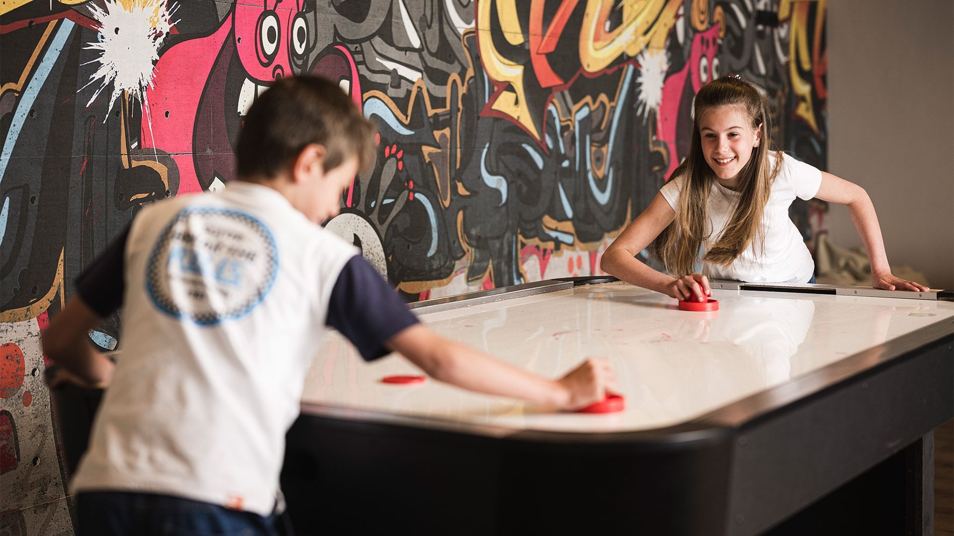Zwei Kinder spielen Air Hockey vor einer bunten Graffiti-Wand.