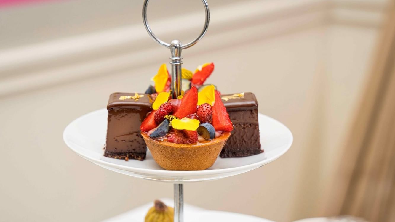Three-tier tray of desserts including tarts, macarons, and sandwiches on a white table.