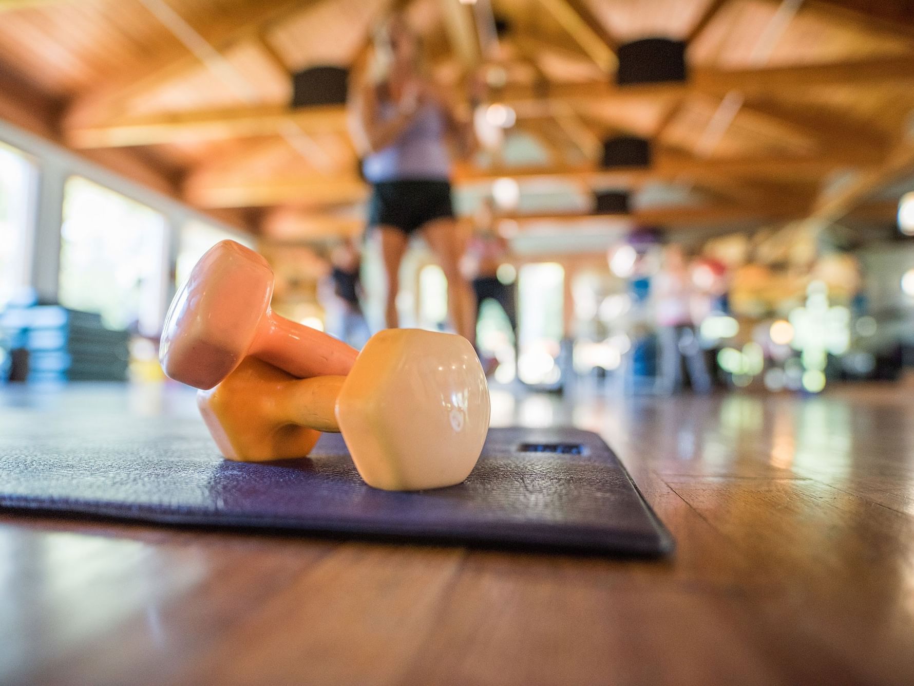 Gym interior with two dumbbells on a mat, and people exercising in the background.