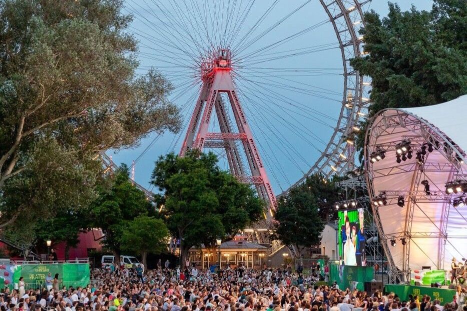 Besucher genießen den Kultursommer Wien im Prater mit Bühne, Live-Performance und dem Wiener Riesenrad im Hintergrund.