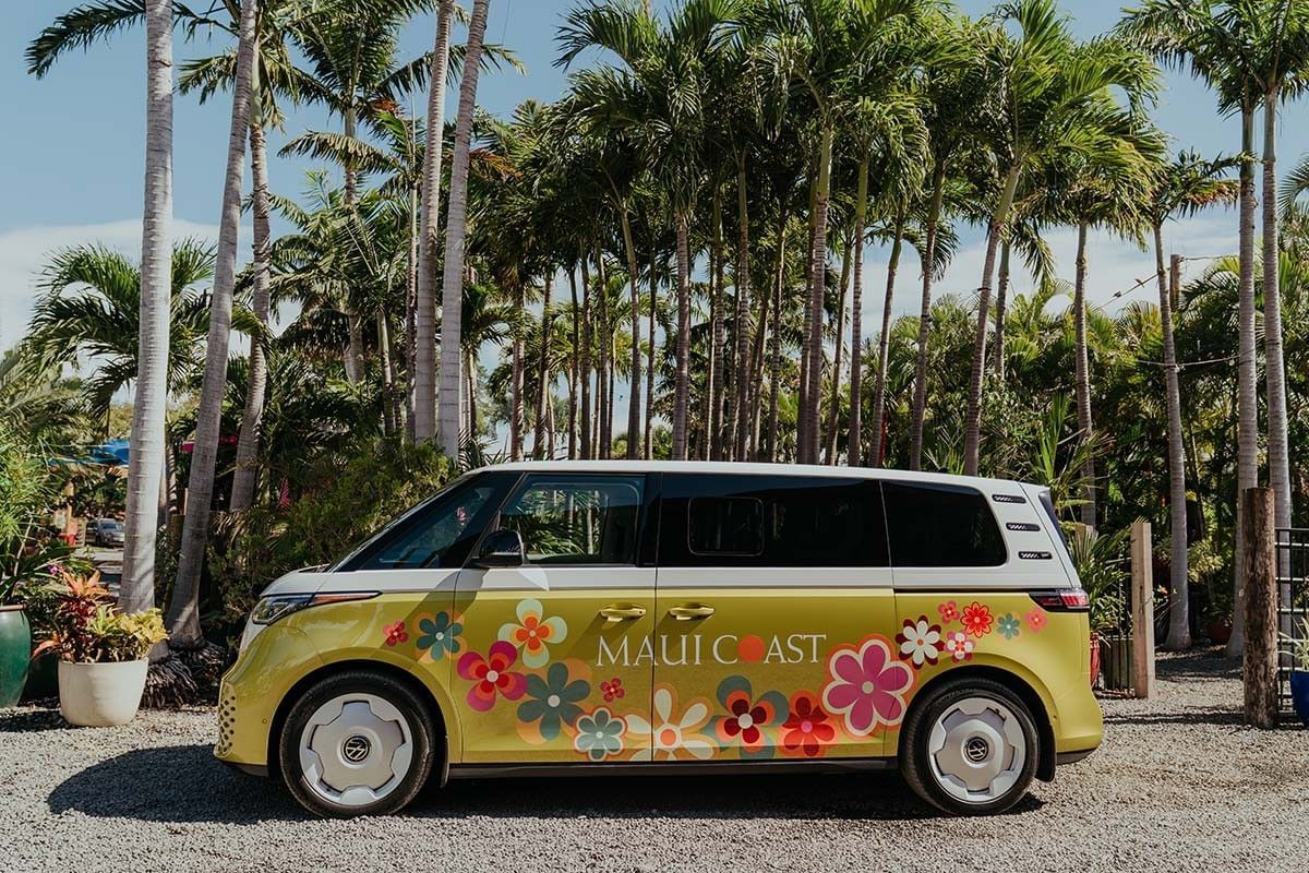 A yellow and white van with colorful flowers and the words 'Maui Coast' parked on gravel in front of palm trees.