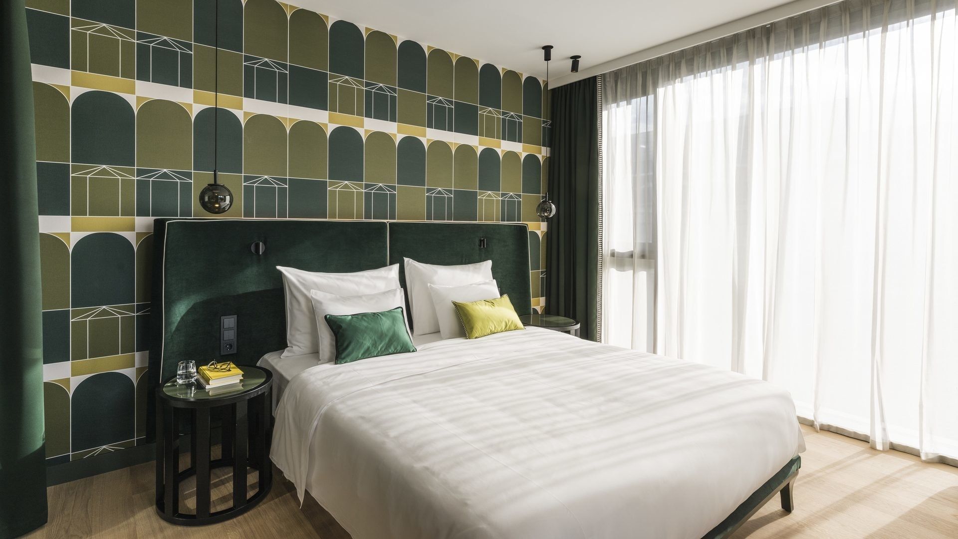 A bedroom with a white bed, green pillows, and a patterned wall, illuminated by a window with curtains.