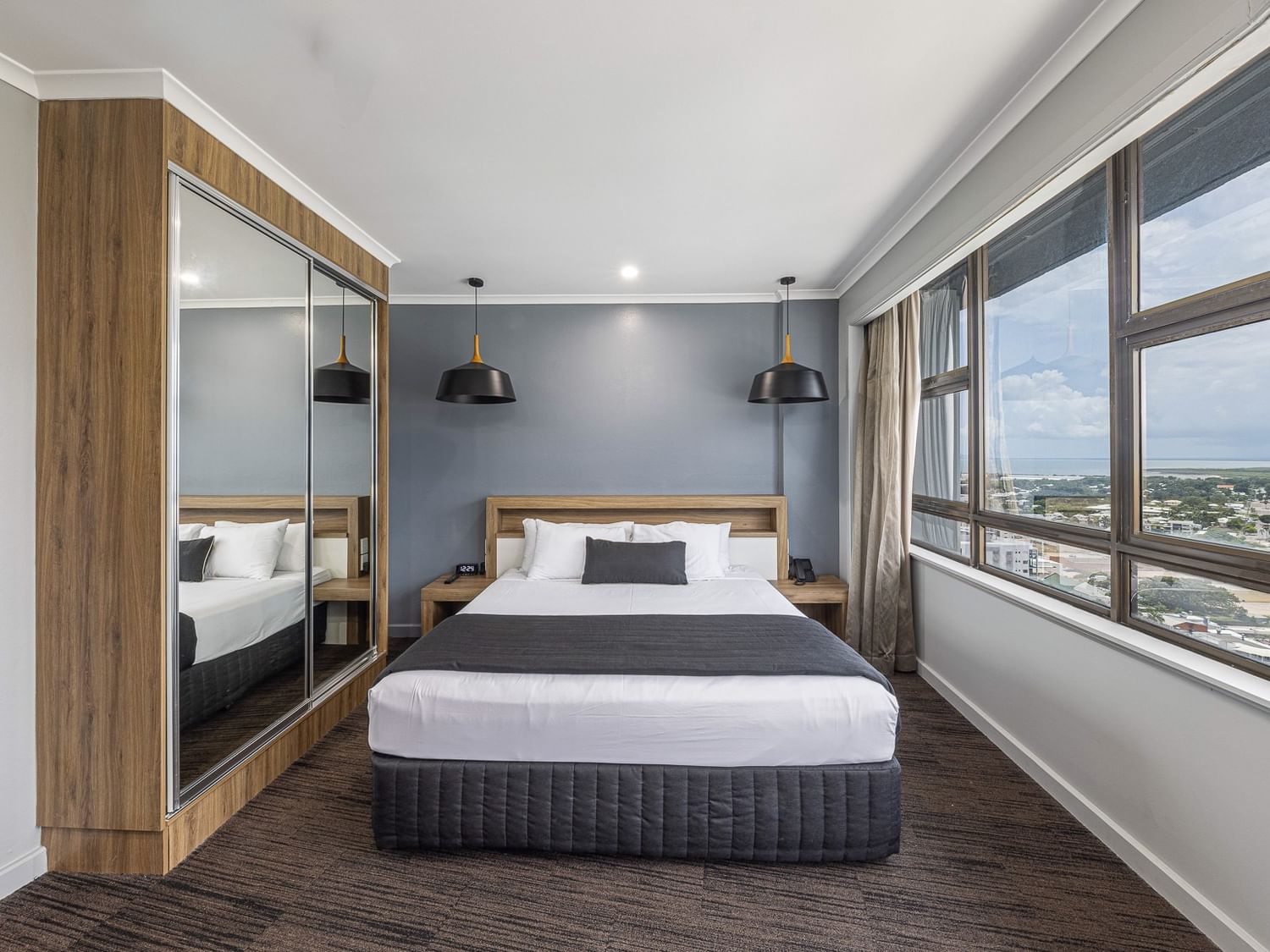 odern executive suite with a king bed, grey accent pillows, black pendant lights, and mirrored wardrobes. Large windows reveal a coastal city view.