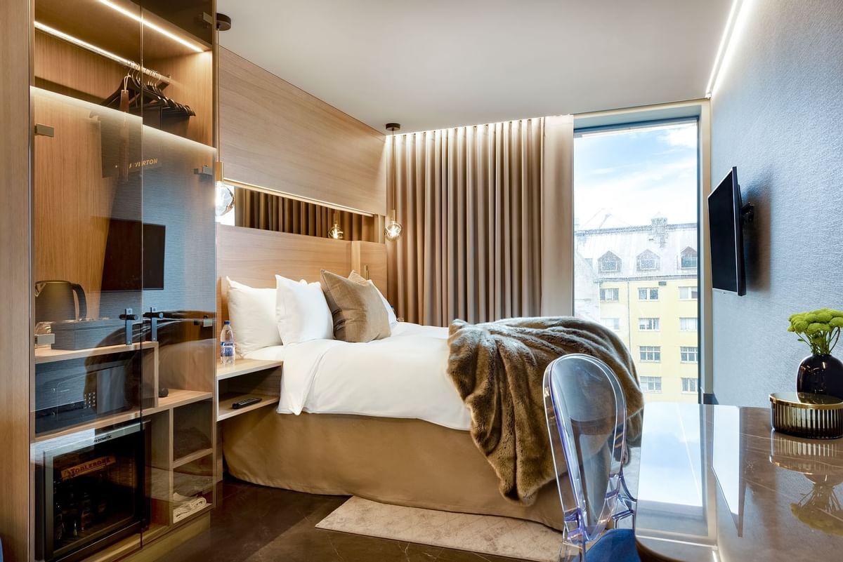 Hotellrum med säng, garderob, bord, stol och utsikt över byggnad och himmel.