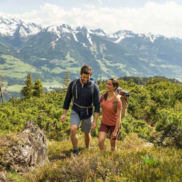 Paar mit Rucksäcken wandert auf einem Bergpfad mit Schneebergen im Hintergrund.