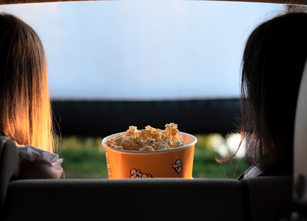 The silhouette of two people sitting in front of an outdoor movie screen with a bucket of popcorn between them. 