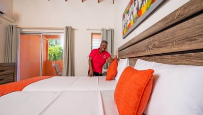 Smiling housekeeper adjusting pillows on a large comfortable bed with orange accents at Worthing Court Apartment