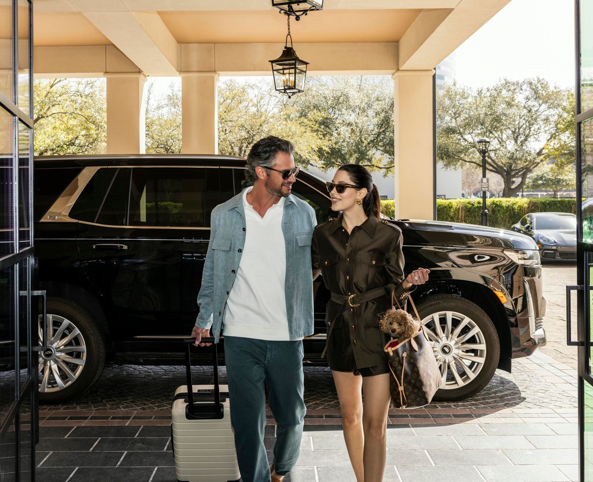 Couple walking toward a luxury SUV with luggage and a small dog at the stylish entrance of Granduca Houston