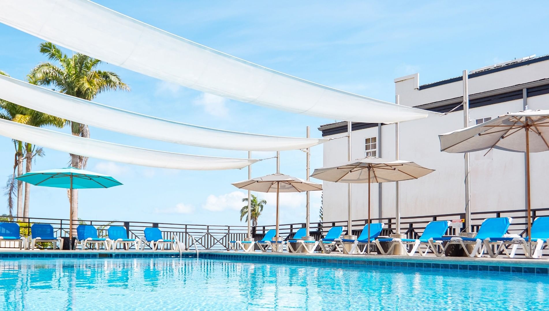 Bright outdoor swimming pool area with blue lounge chairs, tan umbrellas, and large white shade canopies at Bel Jou Hotel