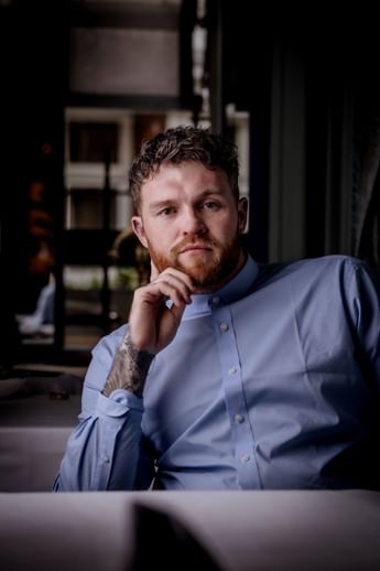 Chef Tom Brown resting his hand on his chin while sitting in a light blue shirt in Tom Brown at The Capital