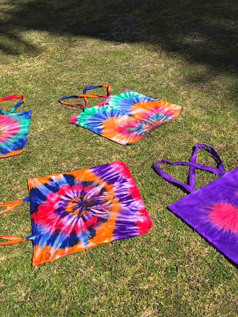 Four colorful tie-dye tote bags displayed on grass showcasing vibrant spiral patterns in rainbow and purple hues.