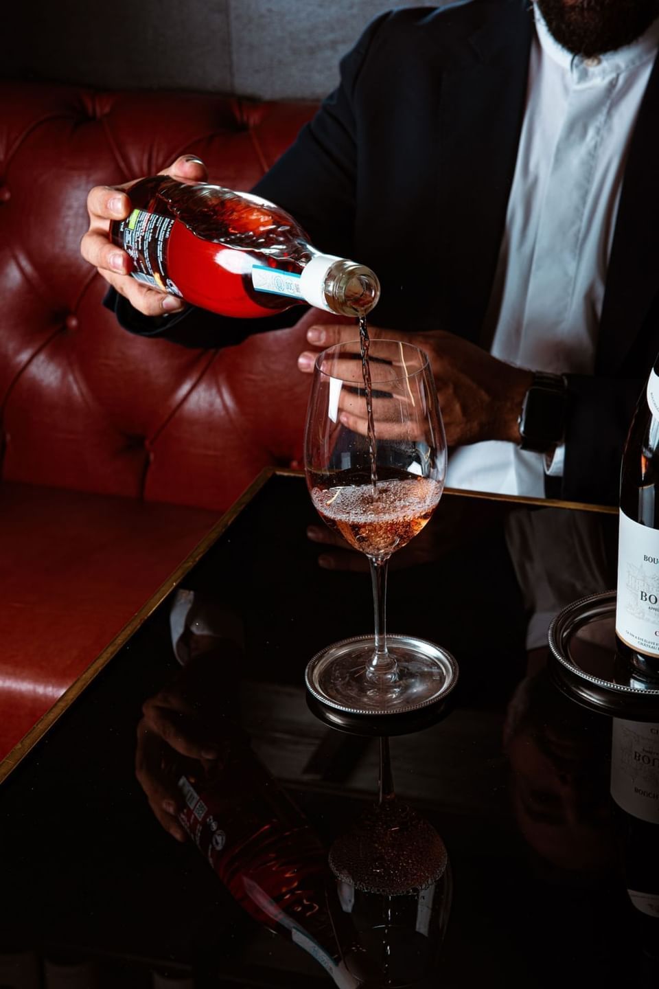 A man pouring rosé wine into a glass on a polished table, with a wine bottle reflection and a red leather backdrop