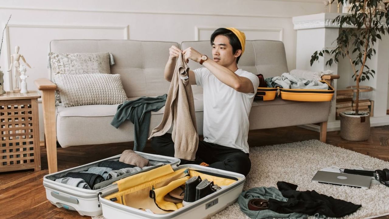 Person sitting on the floor of a living room, packing clothes into a suitcase with folded items and shoes neatly arranged.