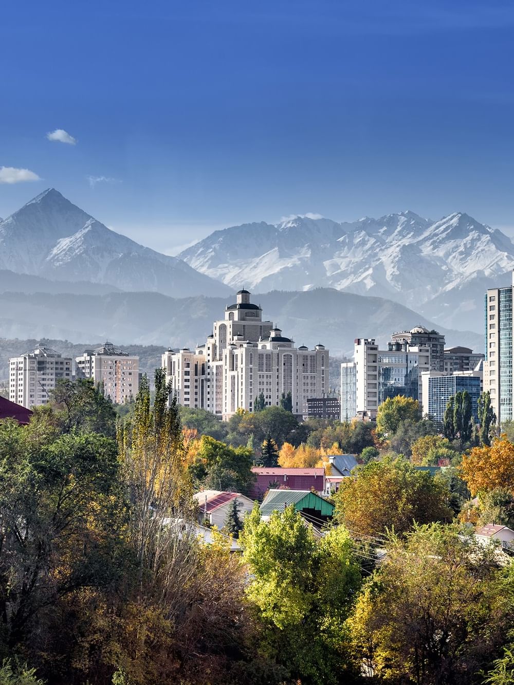 Panoramic view of Almaty cityscape with modern high-rise buildings and autumn foliage against snow-capped mountain range.