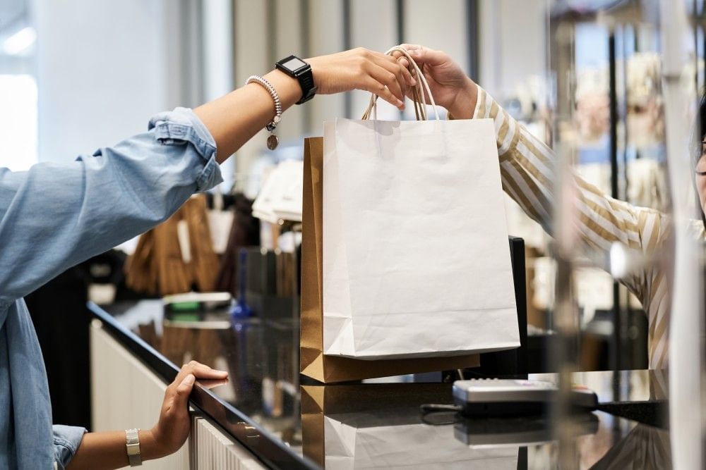 A close-up of a retail worker handing a paper shopping bag to a female customer over a store counter.