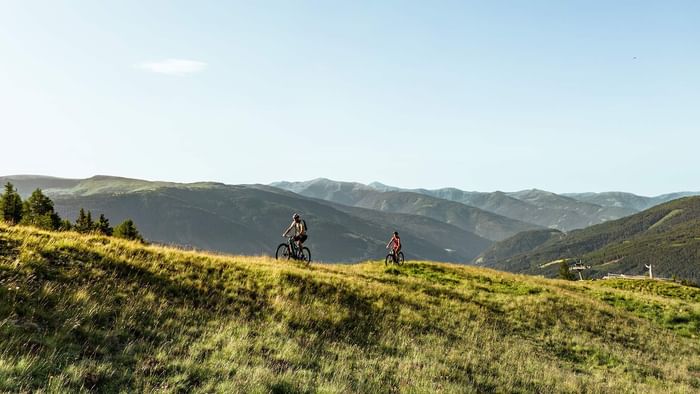 Zwei Radfahrer auf einem Berghang mit Blick auf eine Bergkette im Hintergrund.