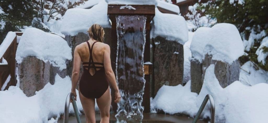 A woman in a swimsuit steps into an outdoor hot pool surrounded by snow, with a waterfall feature flowing behind her.