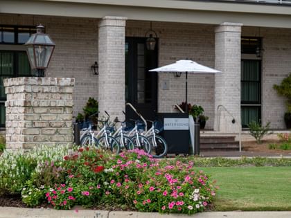 Brick building exterior with colorful flower beds, bike rental station, and outdoor seating area with white umbrella.