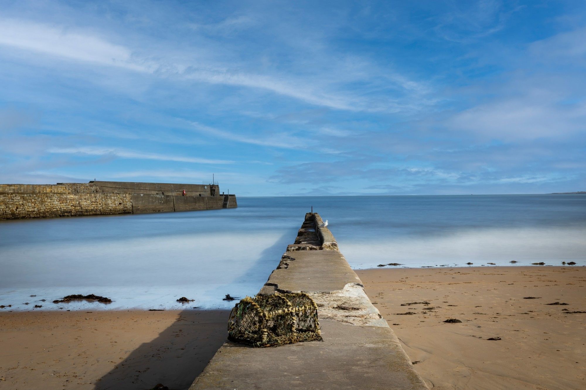 Long concrete pier with a rusted lobster trap on it, leading out to calm seas near Seaton House