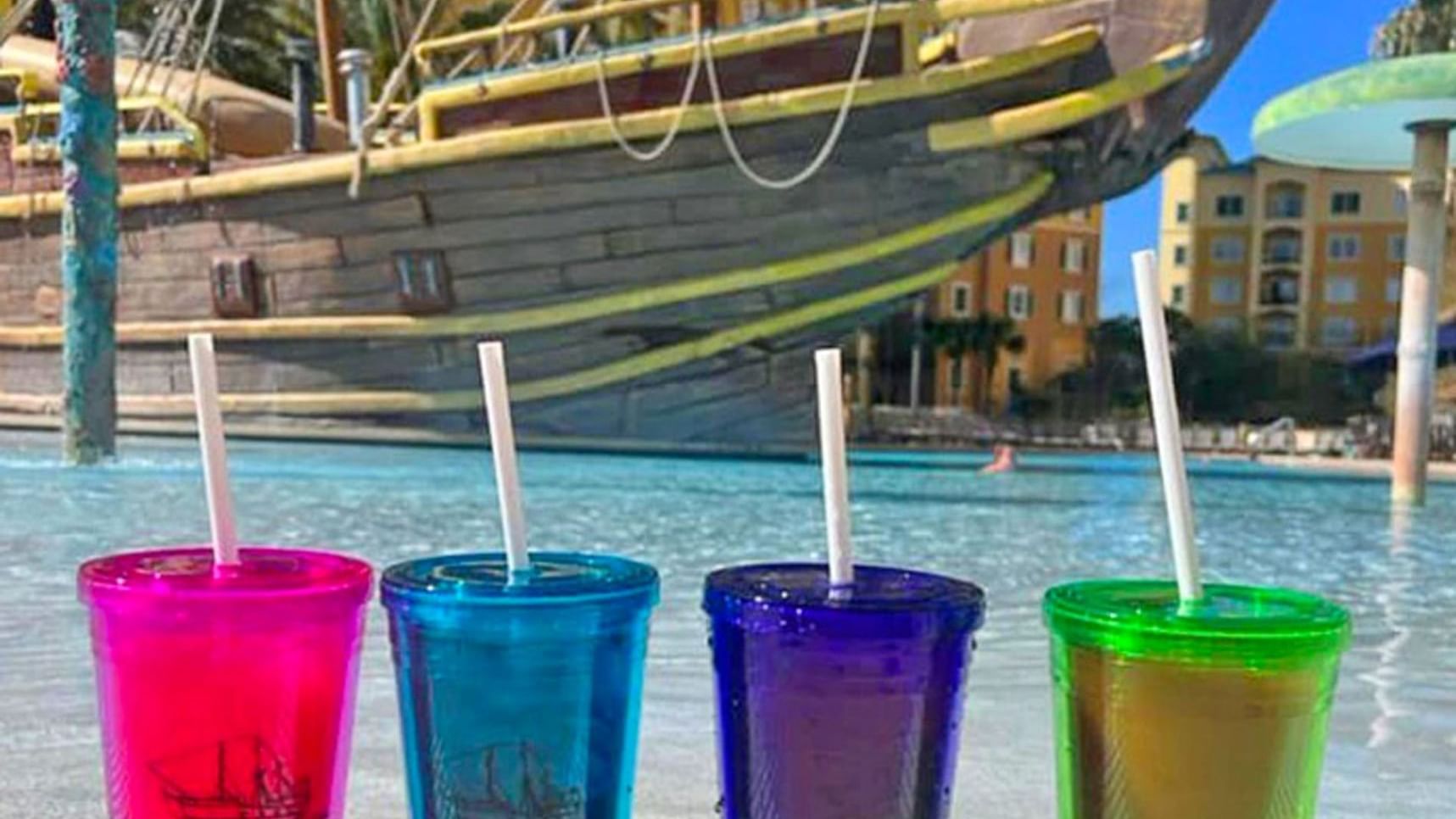 Four colorful cups with straws in front of a pool with a ship in the background.
