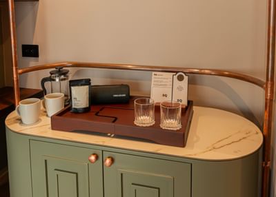 Glassware on a wooden tray by a copper rail under a warm glow on a green cabinet at 5Q House Chapinero