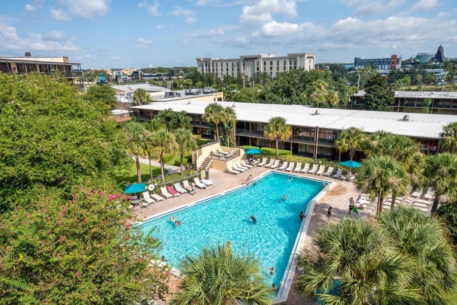 An aerial shot of a two-story hotel building beside a clear blue swimming pool, surrounded by green trees and distant buildings. 