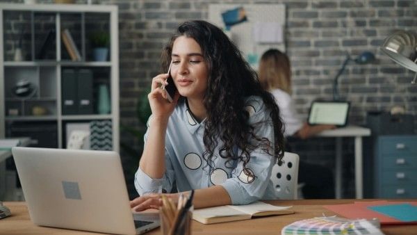 A woman making a phone call inside a lounge while looking at a laptop.