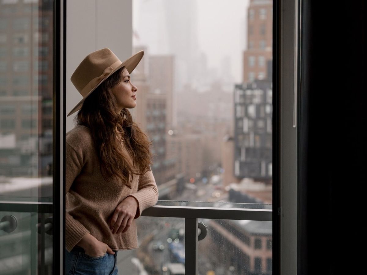 Woman in hat leaning on balcony railing and overlooking foggy city street in Manhattan Suite at Gansevoort Meatpacking NYC