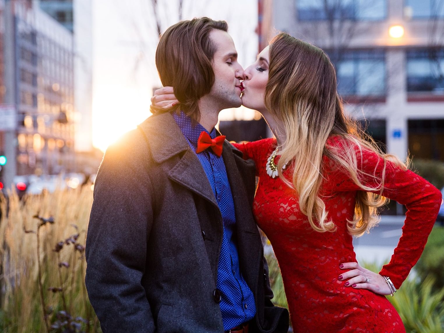 Couple kissing outdoors in urban setting with buildings and greenery, man in bow tie, woman in red dress.