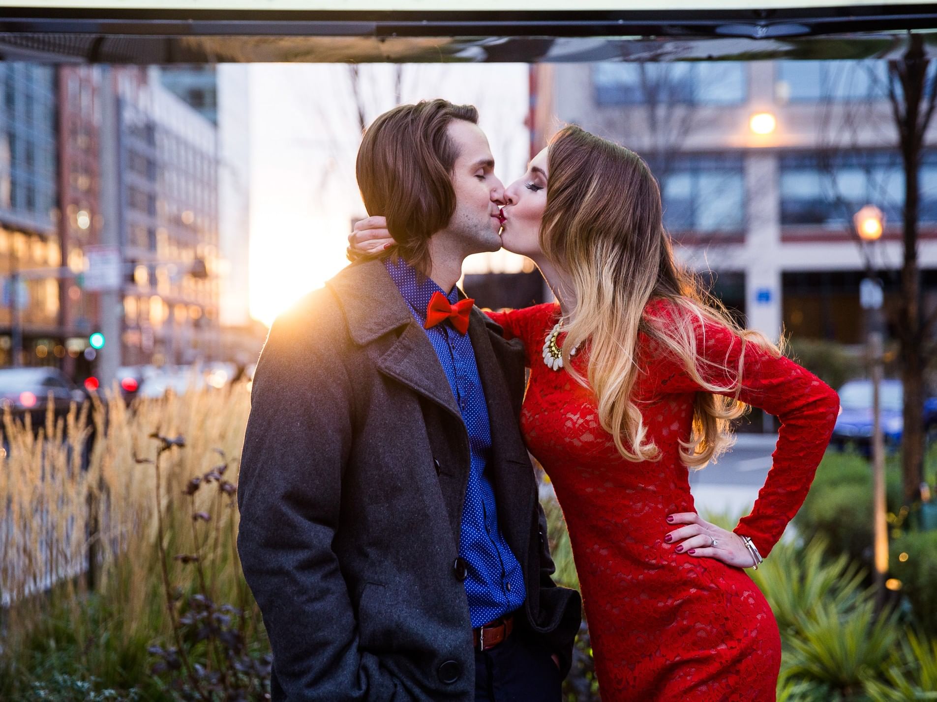 Couple kissing outdoors in urban setting with buildings and greenery, man in bow tie, woman in red dress.