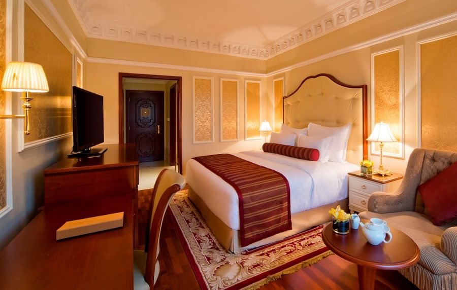 Tea set on a table by an armchair under a crown molding ceiling near a king-sized bed in Deluxe Room at Warwick Doha Hotel
