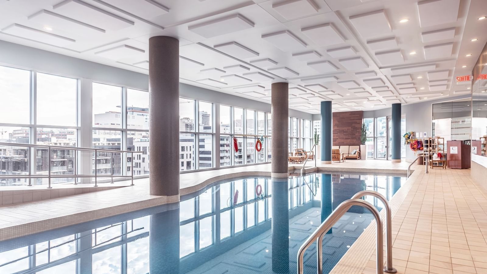 Indoor swimming pool with lounge chairs by large windows under a tiled ceiling at Warwick Le Crystal Montreal