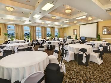 A large meeting room with round tables, white tablecloths, and black chairs with white ribbons, featuring a projector screen and windows with curtains.