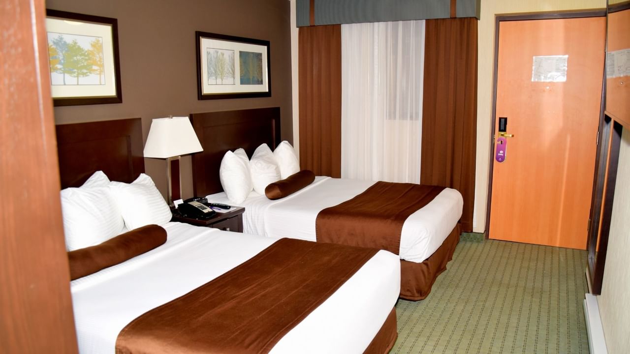 Two beds, lamp, telephone, framed pictures, curtains, and wooden door in a hotel room.