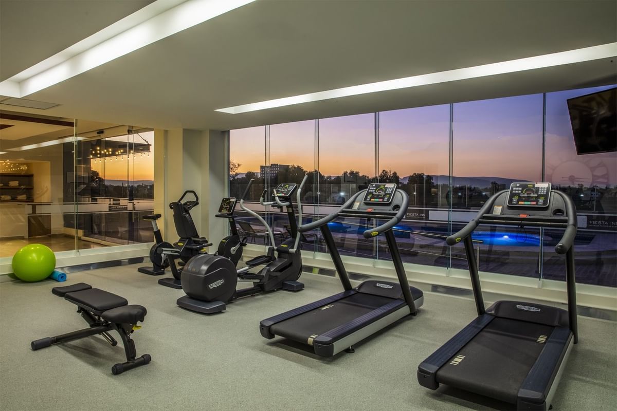 Gimnasio equipado con máquinas de cardio y vista a la terraza de la piscina al atardecer en Fiesta Inn