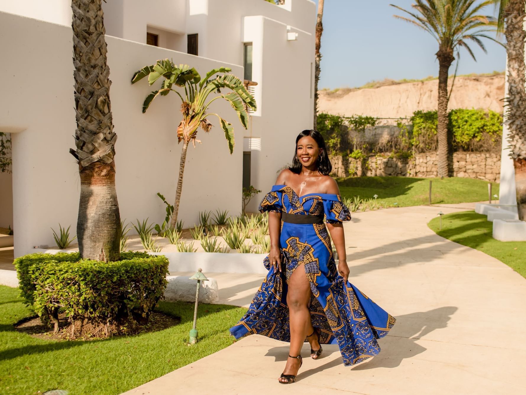 A stylish woman in a vibrant blue and gold off-the-shoulder dress, walks along a path at Marquis Los Cabos Resort