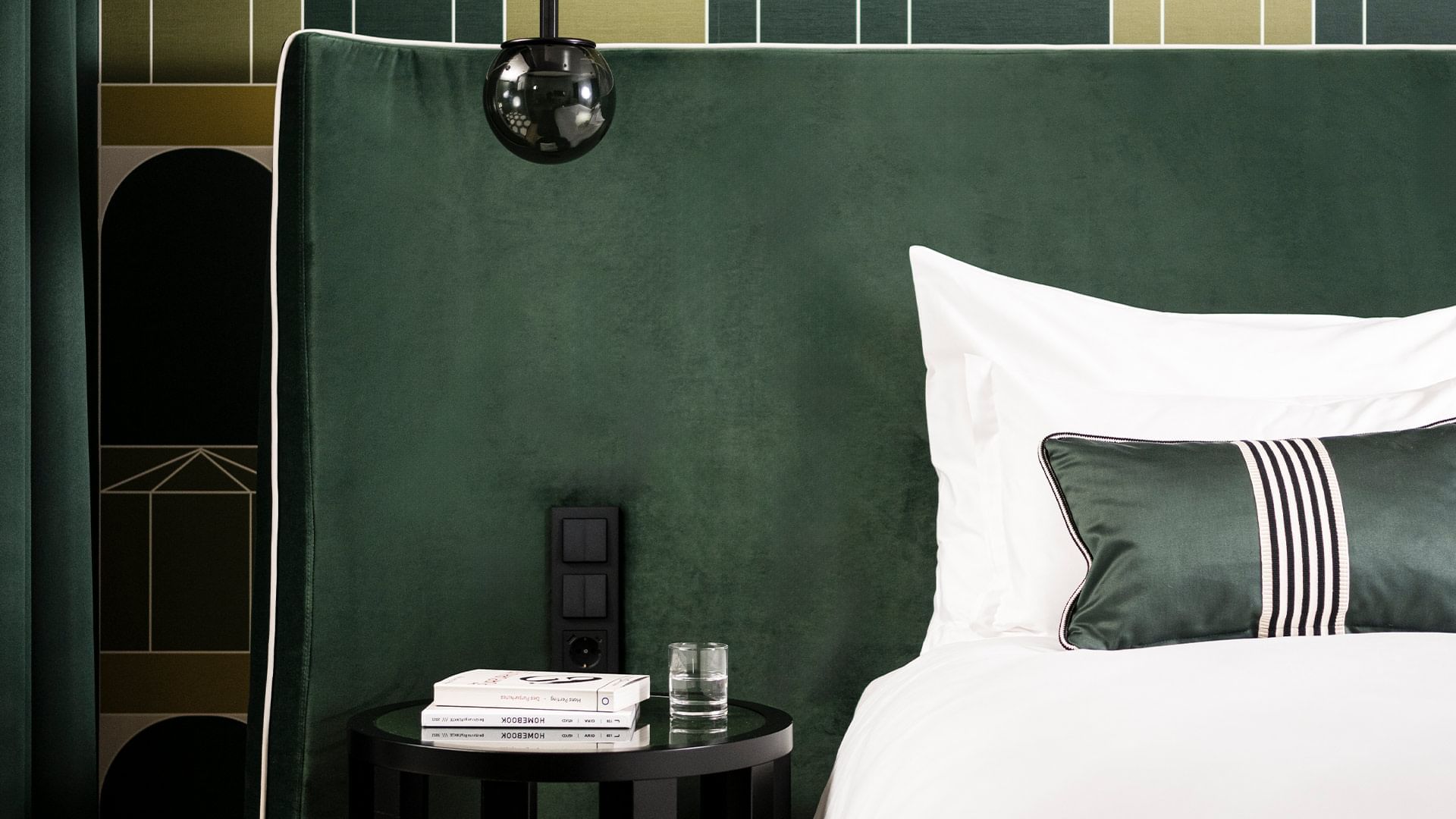 Zimmer mit grünem Bett, Kissen, Nachttisch mit Glas und Buch, und schwarzer Lampe.