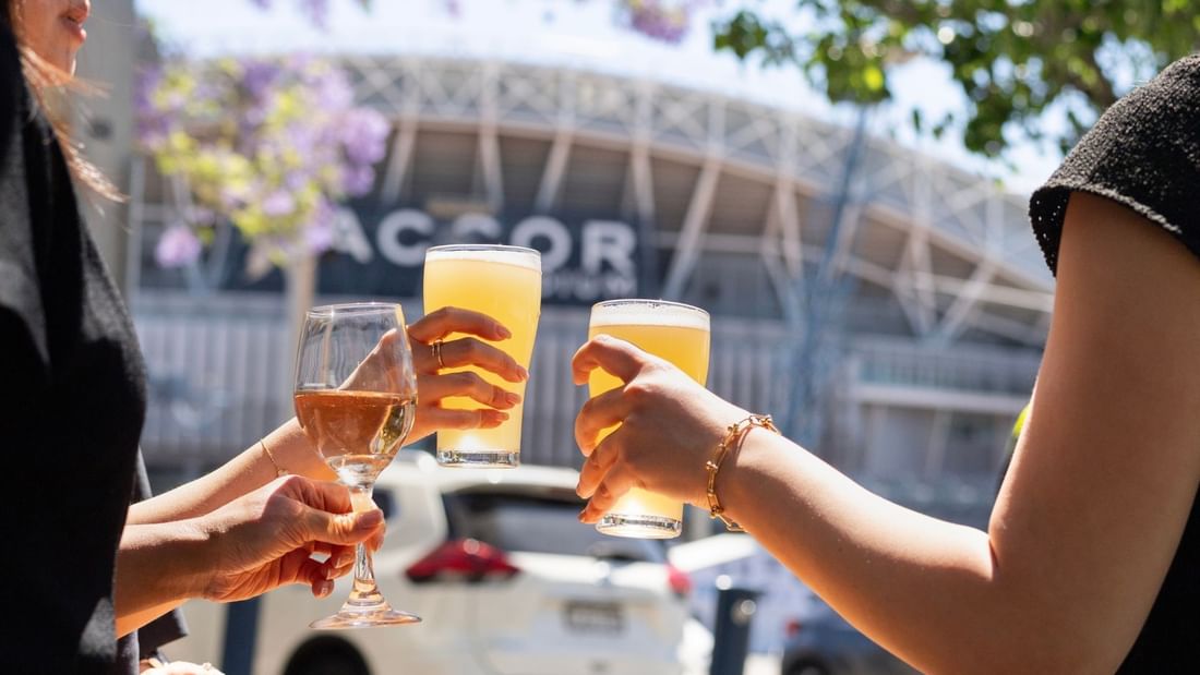 Three people toasting with wine and beer glasses outdoors at The Brewery Bar + Kitchen at Novotel Sydney Olympic Park.
