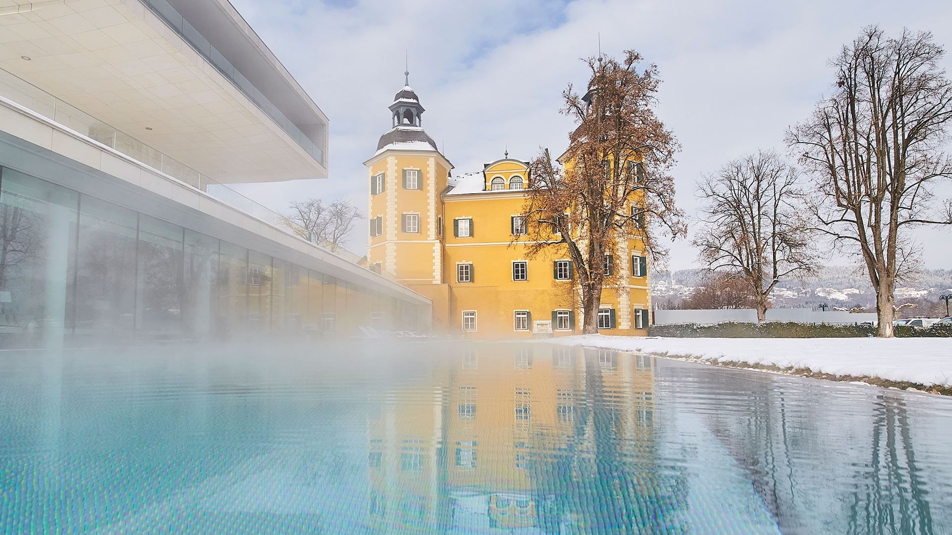Yellow castle reflected in pool water with modern building and winter landscape at Falkensteiner Schlosshotel Velden