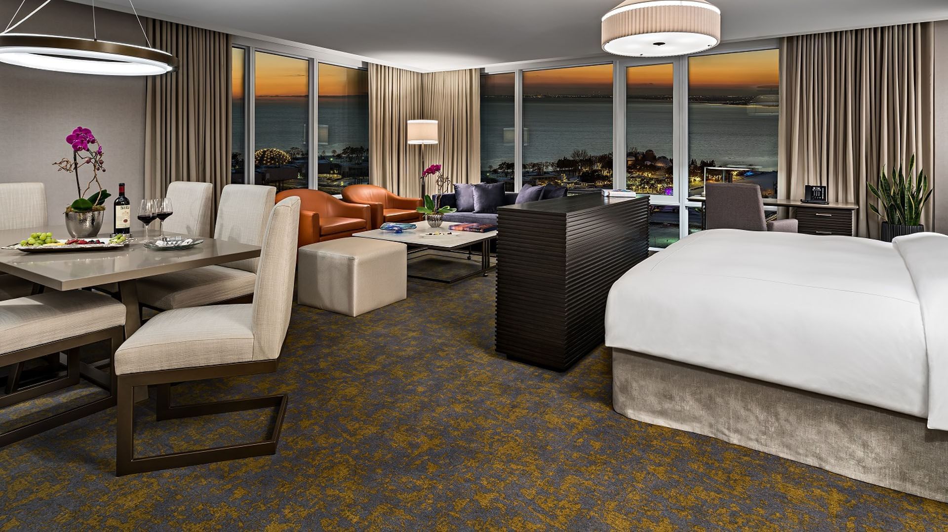 Spacious hotel suite with plush bed and dining area facing floor-to-ceiling sunset ocean views at Hotel X Toronto